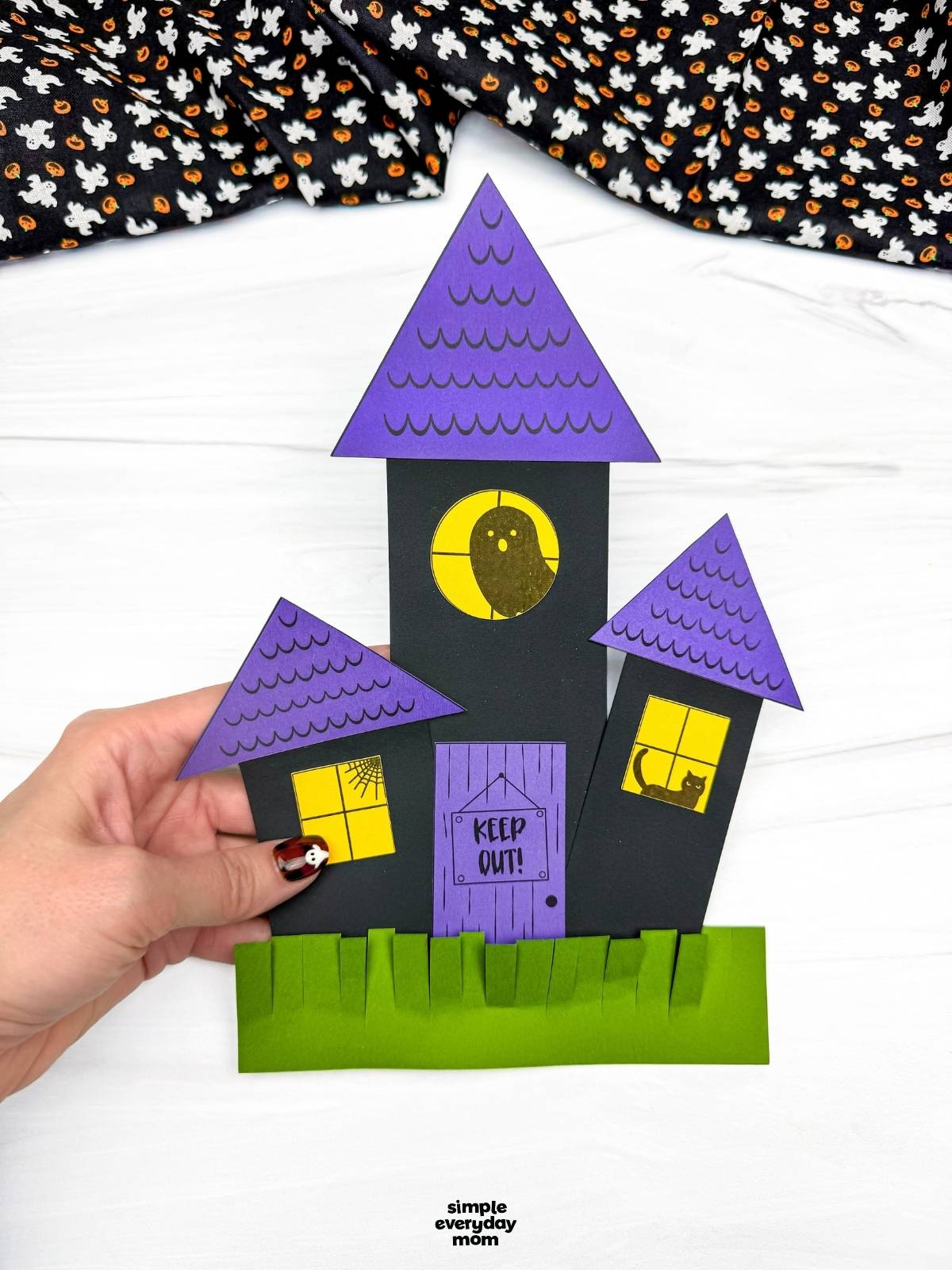 Hands holding a completed haunted house shape craft with triangular roofs, windows, a door, and fringed grass.