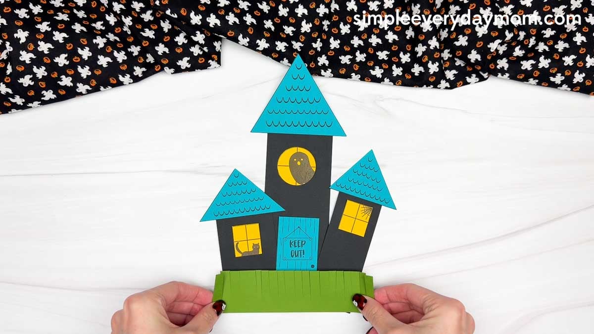 Hands holding a completed haunted house shape craft with triangular roofs, windows, a door, and fringed grass.