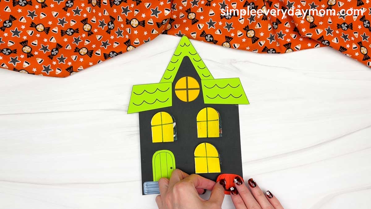 Hands attaching the pumpkin to the bottom right of the Peekaboo Haunted House Craft.