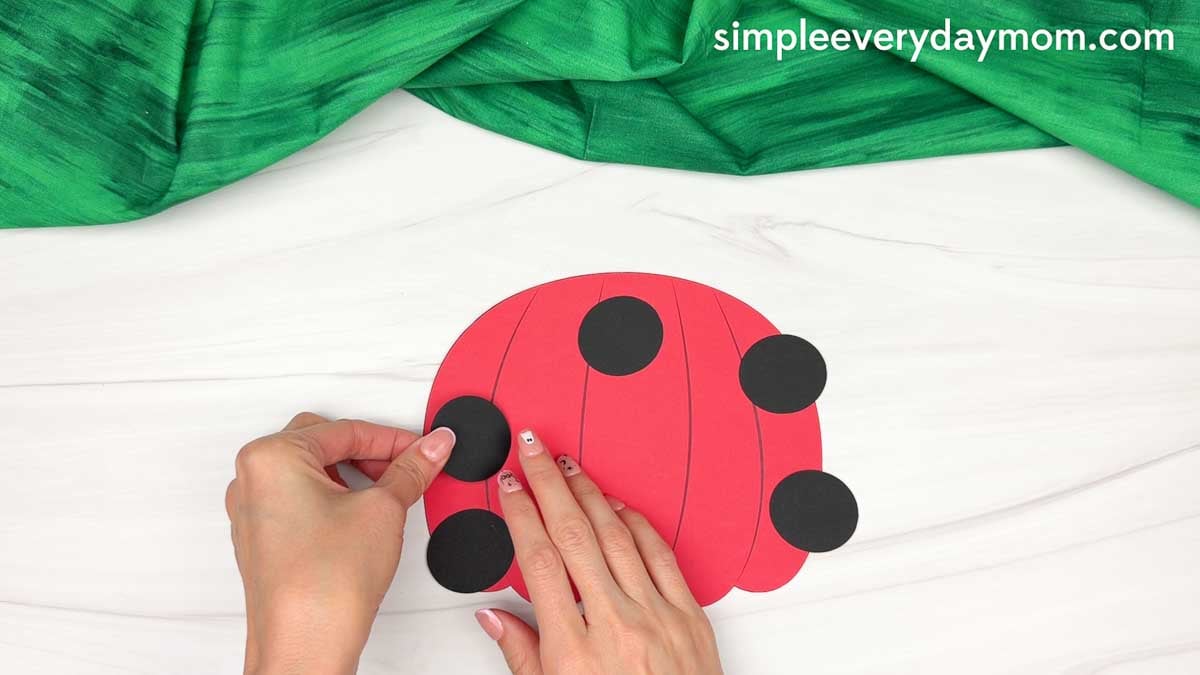 Hands gluing black spots onto the red pumpkin body for the ladybug pumpkin craft.