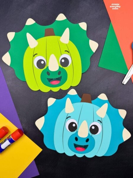 Side by side completed green and blue dinosaur pumpkin disguise crafts with horns and spiked frills beside colorful craft supplies.