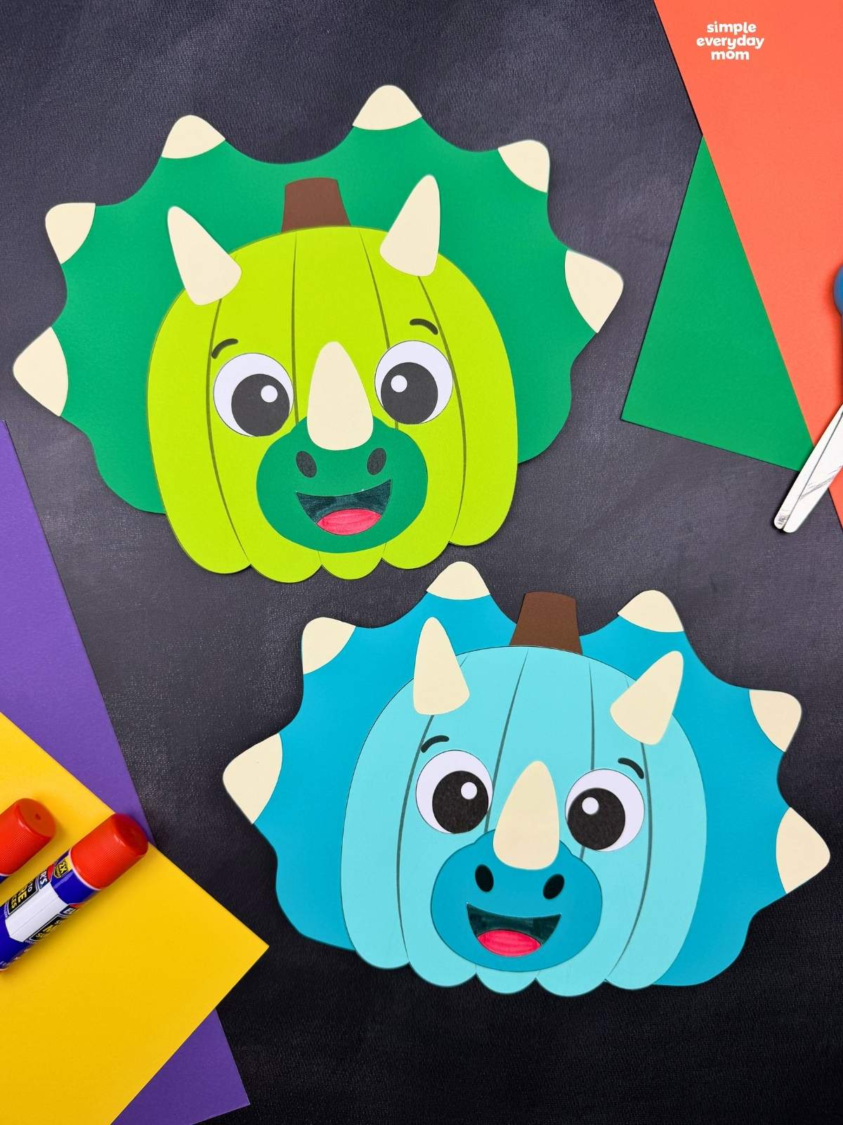 Side by side completed green and blue dinosaur pumpkin disguise crafts with horns and spiked frills beside colorful craft supplies.