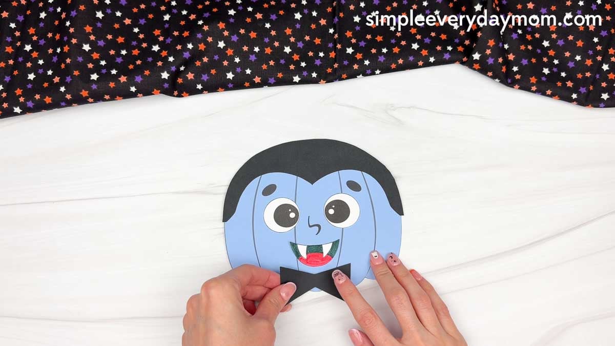 Hands placing the bow tie at the bottom center of the vampire pumpkin face.