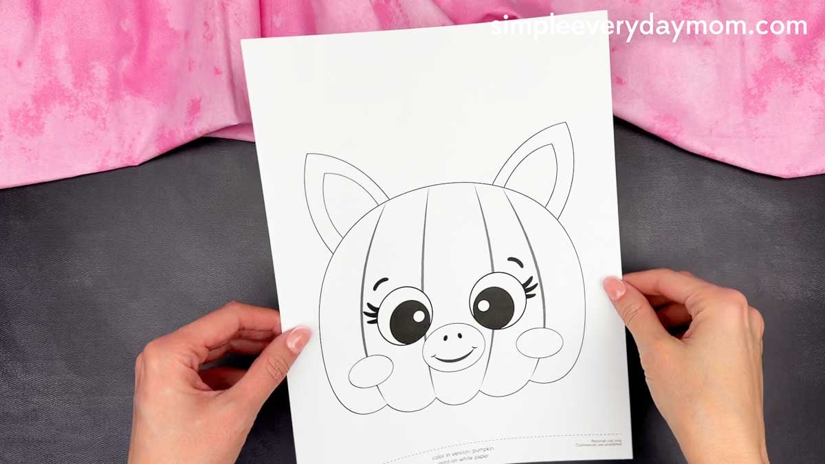 Hands holding a printable unicorn pumpkin craft template featuring a pumpkin face with ears, eyes, cheeks, and a smiling mouth.