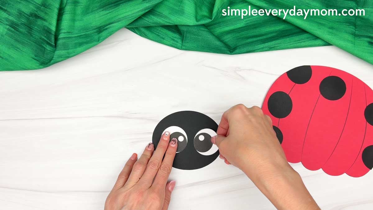 Hands gluing the eyes onto the ladybug head beside the spotted pumpkin body.