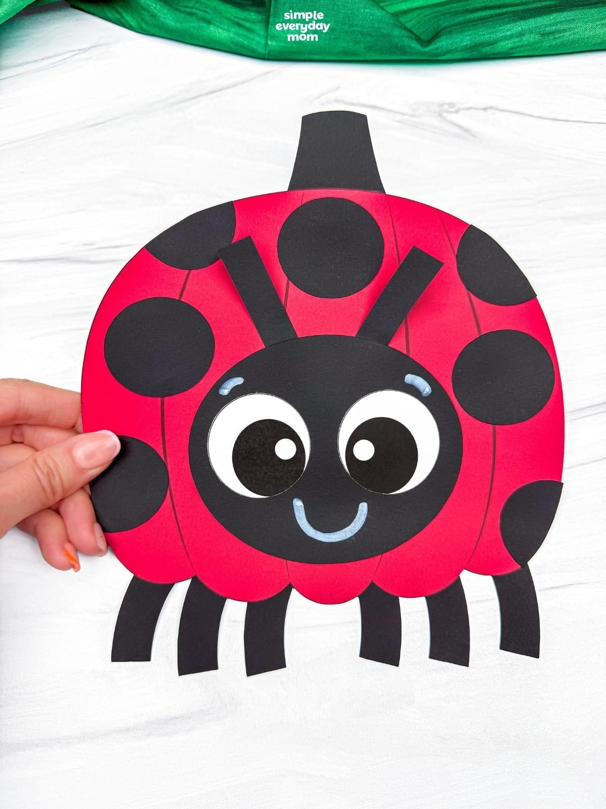 Hands holding a completed ladybug pumpkin craft with black spots, a smiling face, antennae, and six legs.