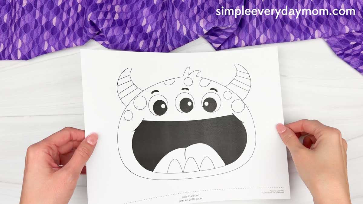 Hands holding a printable monster name craft template with three eyes, striped horns, and a large open mouth.