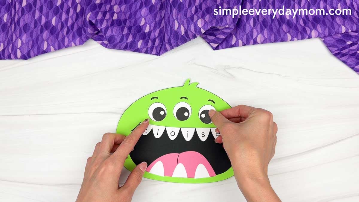 Hands placing an eye onto the monster’s face above the lettered teeth.