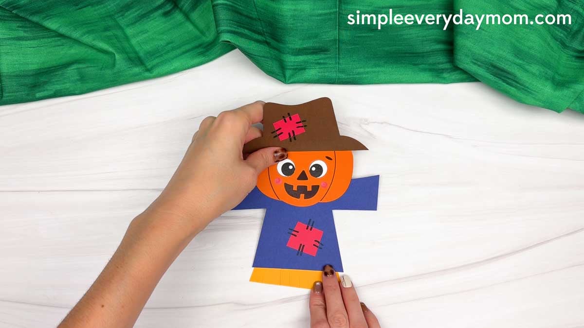 Hands assembling the pumpkin head scarecrow with a patched shirt and straw detail.
