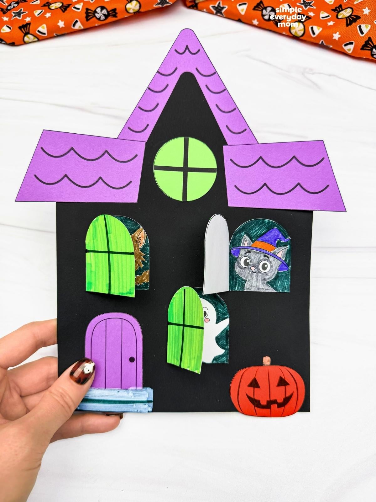 Hands holding a completed Peekaboo Haunted House Craft with open windows revealing Halloween characters, a door, and a pumpkin.