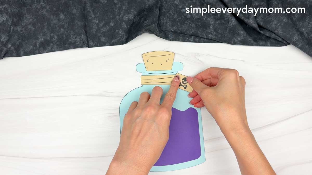 Hands attaching a skull and crossbones tag to the neck of the Halloween potion bottle craft.