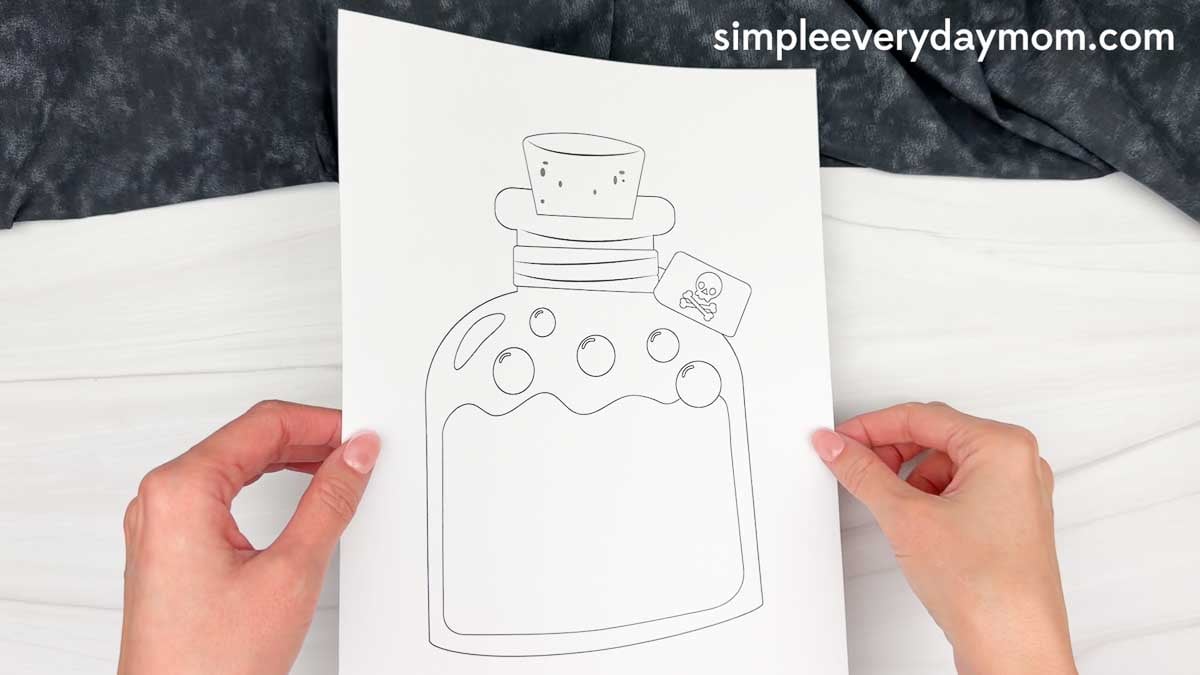 Hands holding a printable Halloween potion bottle coloring page with a cork and skull tag.