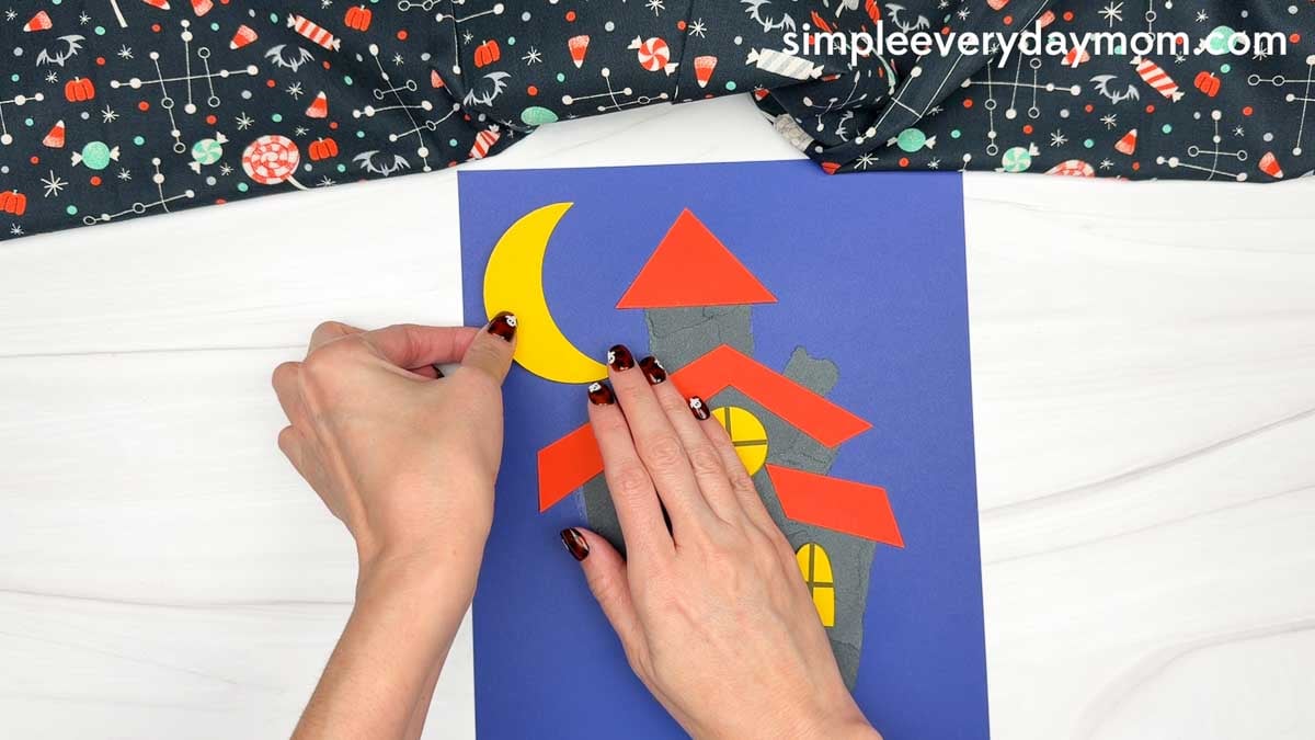 Hands placing a crescent moon beside the torn paper haunted house.