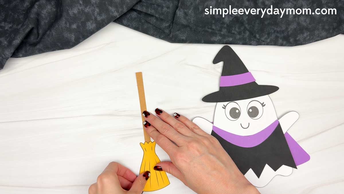 Hands attaching the broom bristles to the handle beside the Witch Ghost Disguise craft.