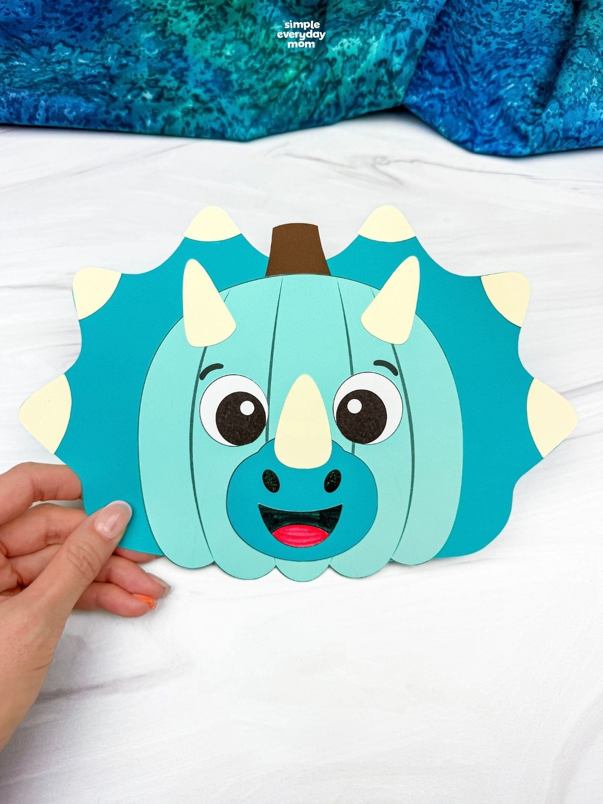 Hands holding a completed blue dinosaur pumpkin disguise craft with horns and a spiked frill.