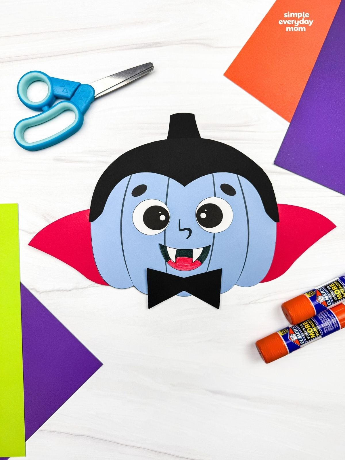 A completed craft showing a vampire pumpkin disguise with a cape, fangs, and bow tie surrounded by craft supplies.