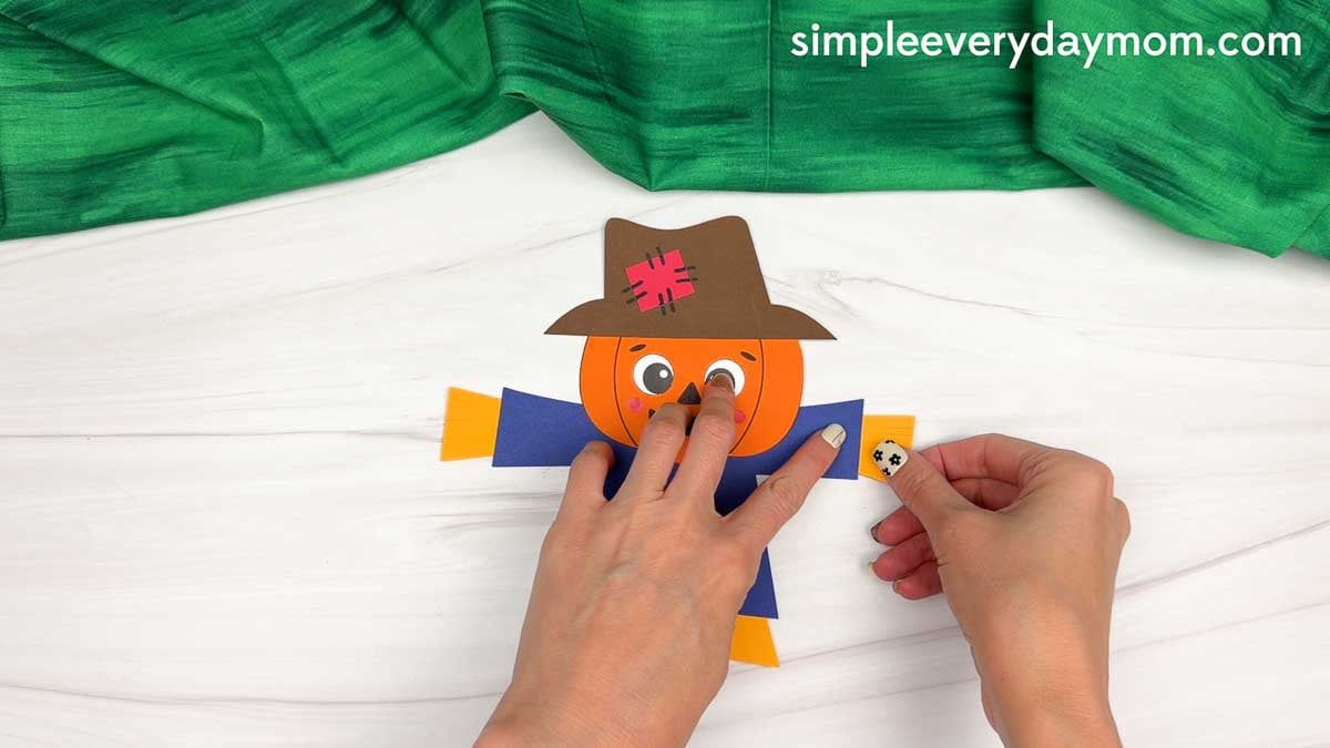 Hands attaching straw pieces to the ends of the scarecrow’s arms.