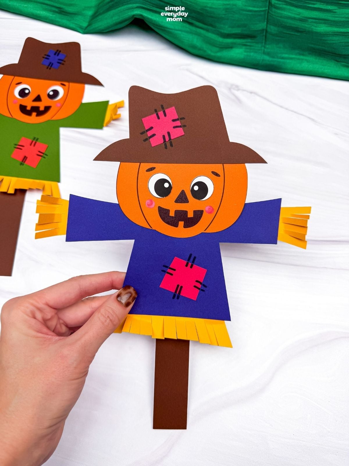 Hands holding a completed pumpkin head scarecrow with a patched hat, shirt, and straw details.