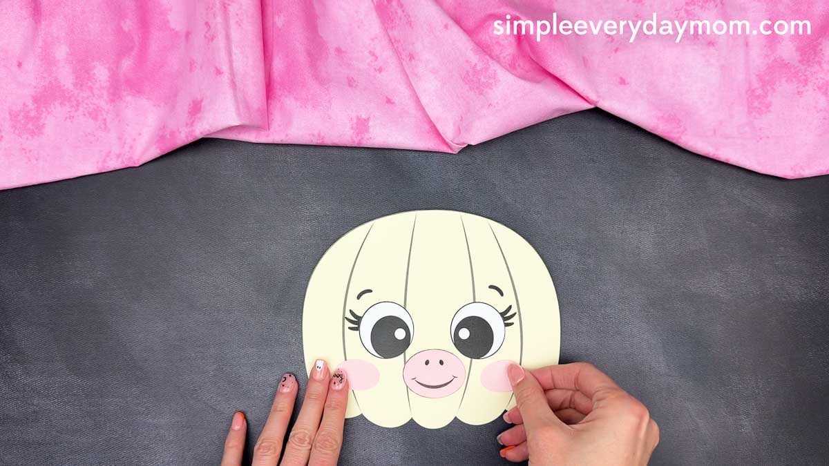 Hands gluing the cheeks on both sides of the unicorn pumpkin craft face.