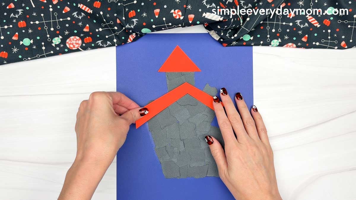 Hands placing the main roof piece onto the torn paper haunted house.