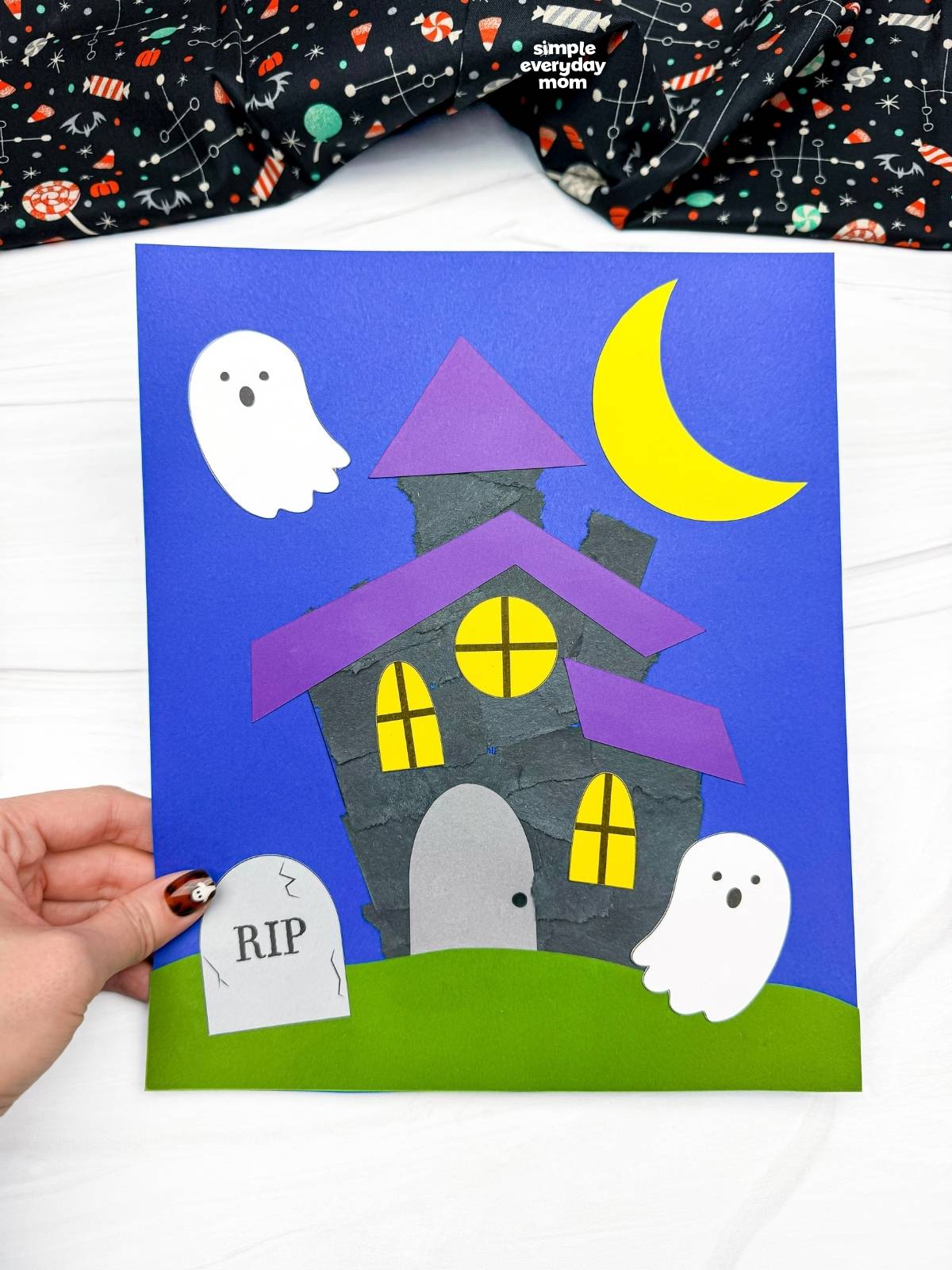 Hands holding a completed Haunted House Torn Paper Craft with ghosts, a crescent moon, windows, grass, and a headstone.