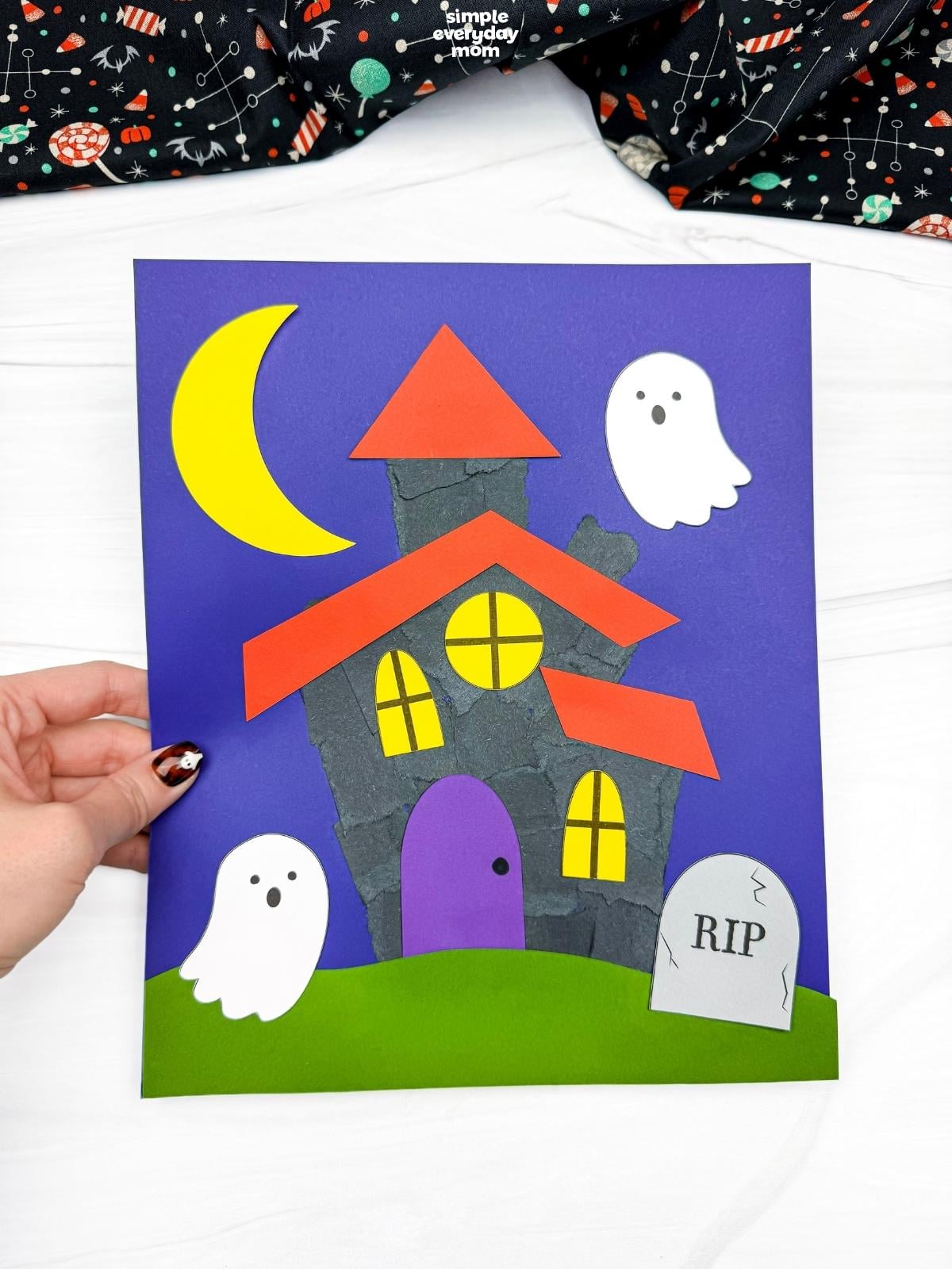 Hands holding a completed Haunted House Torn Paper Craft with ghosts, a crescent moon, windows, grass, and a headstone.
