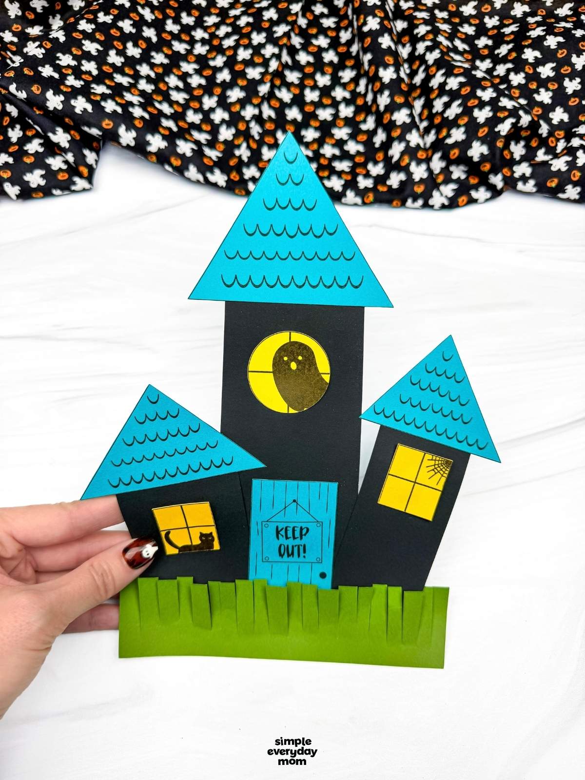 Hands holding a completed haunted house shape craft with triangular roofs, windows, a door, and fringed grass.