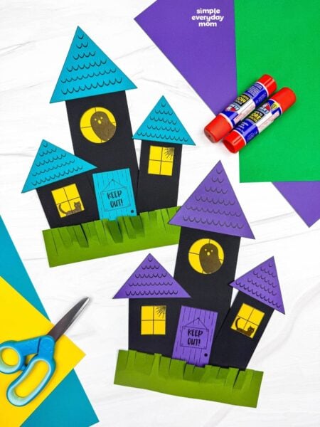Side by side completed haunted house shape crafts with triangular roofs, windows, doors, and fringed grass.