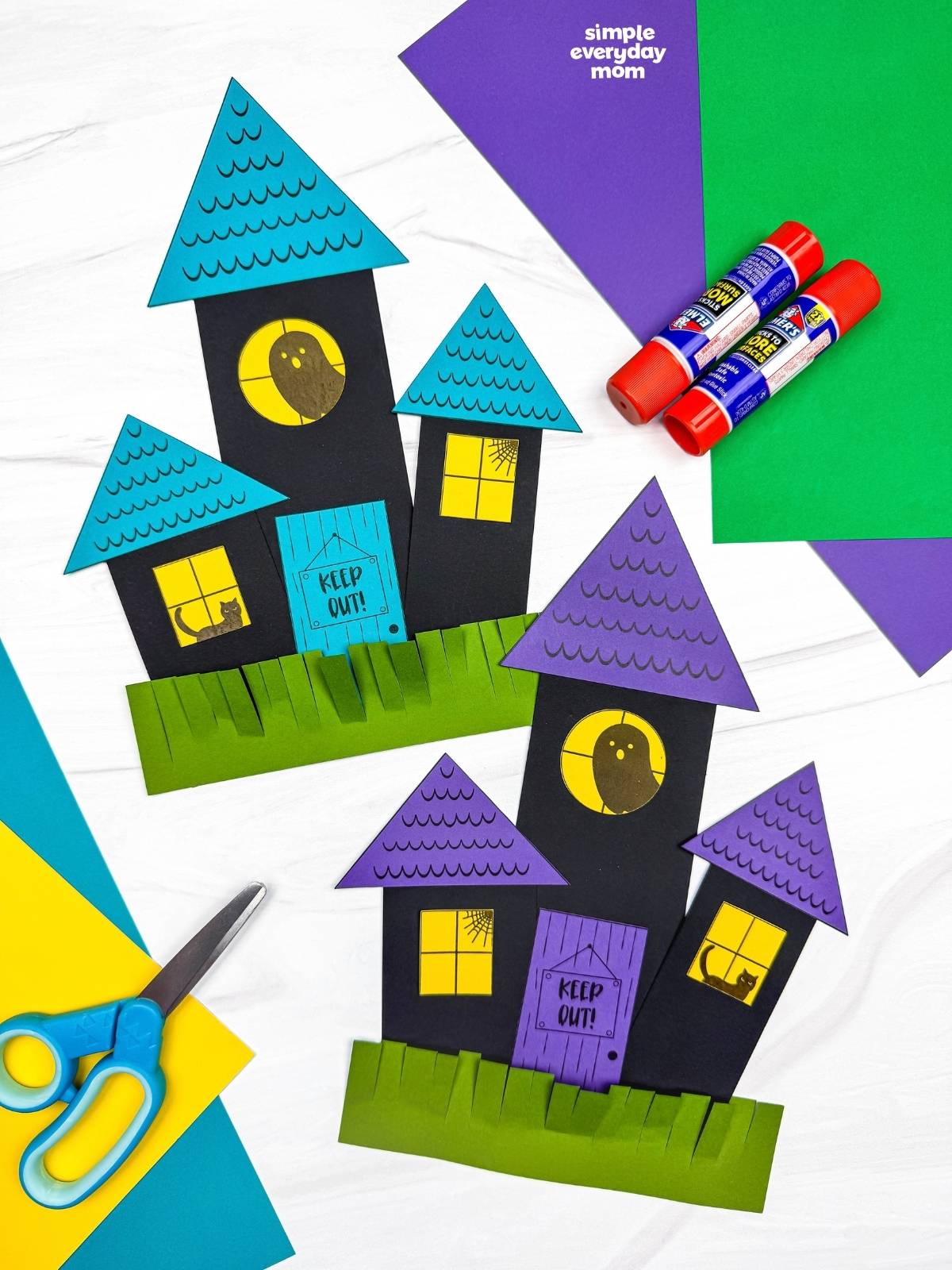 Side by side completed haunted house shape crafts with triangular roofs, windows, doors, and fringed grass.
