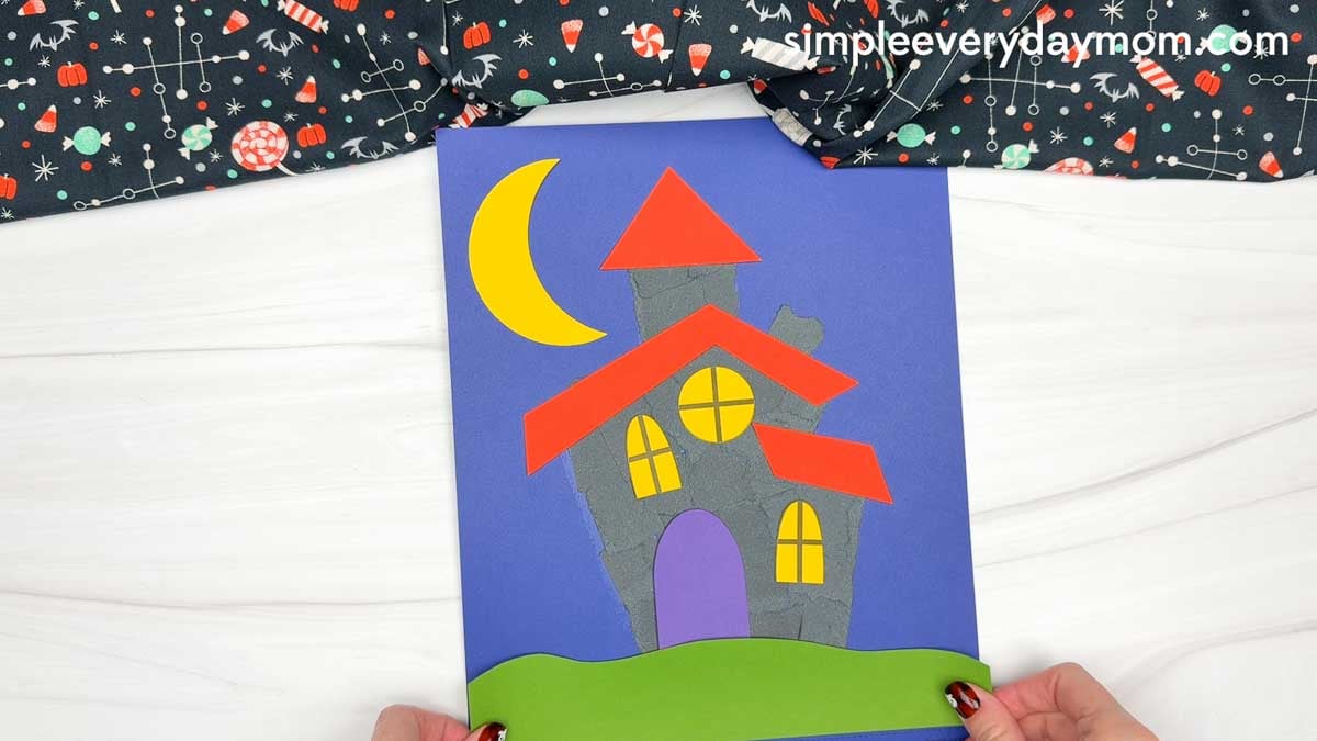 Hands placing the grass piece along the bottom of the torn paper haunted house scene.