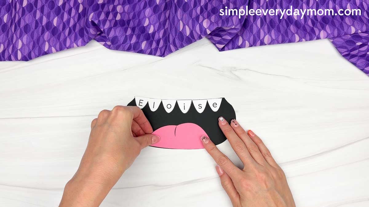 Hands gluing the tongue to the bottom of the monster’s mouth below the lettered teeth.