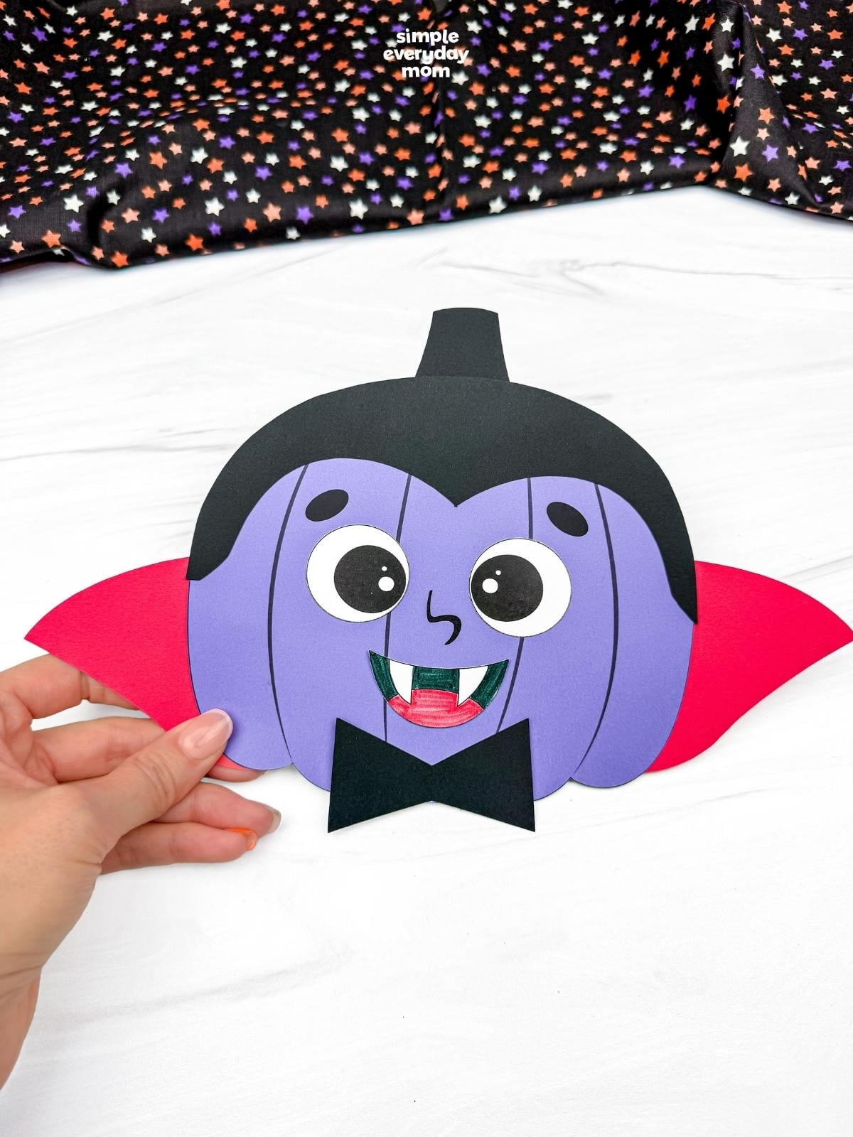 Hands holding a completed vampire pumpkin disguise craft with a cape, fangs, and bow tie.