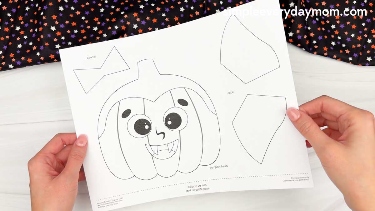 Hands holding a printable vampire pumpkin craft template with a pumpkin head, bow tie, and cape pieces.