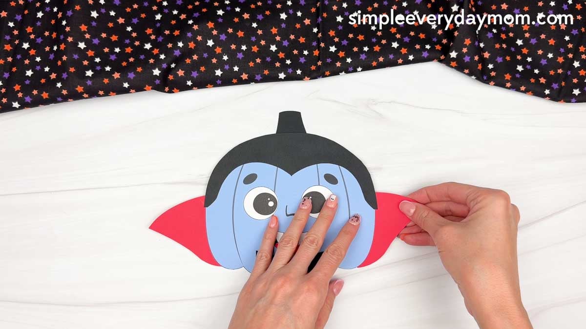 Hands attaching the cape pieces behind both sides of the vampire pumpkin head.