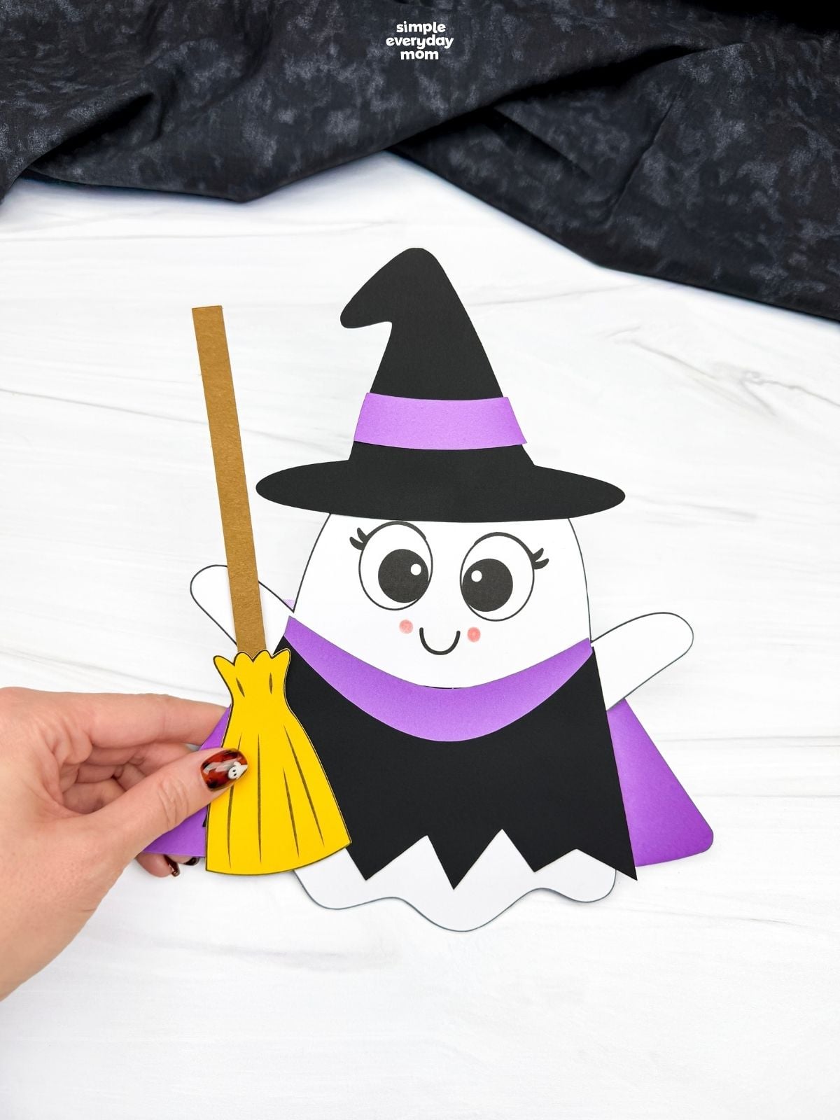 Hands holding a completed Witch Ghost Disguise craft with a witch hat, dress, and broom.