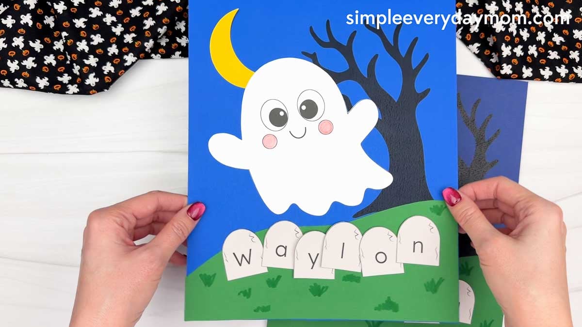 Hands holding a completed Ghost Name Craft featuring a smiling ghost, crescent moon, spooky tree, and tombstones spelling a child's name.