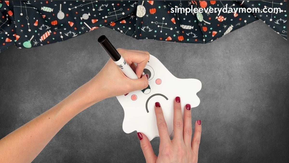 Hands coloring the mouth black on a Halloween paper ghost craft with a marker.