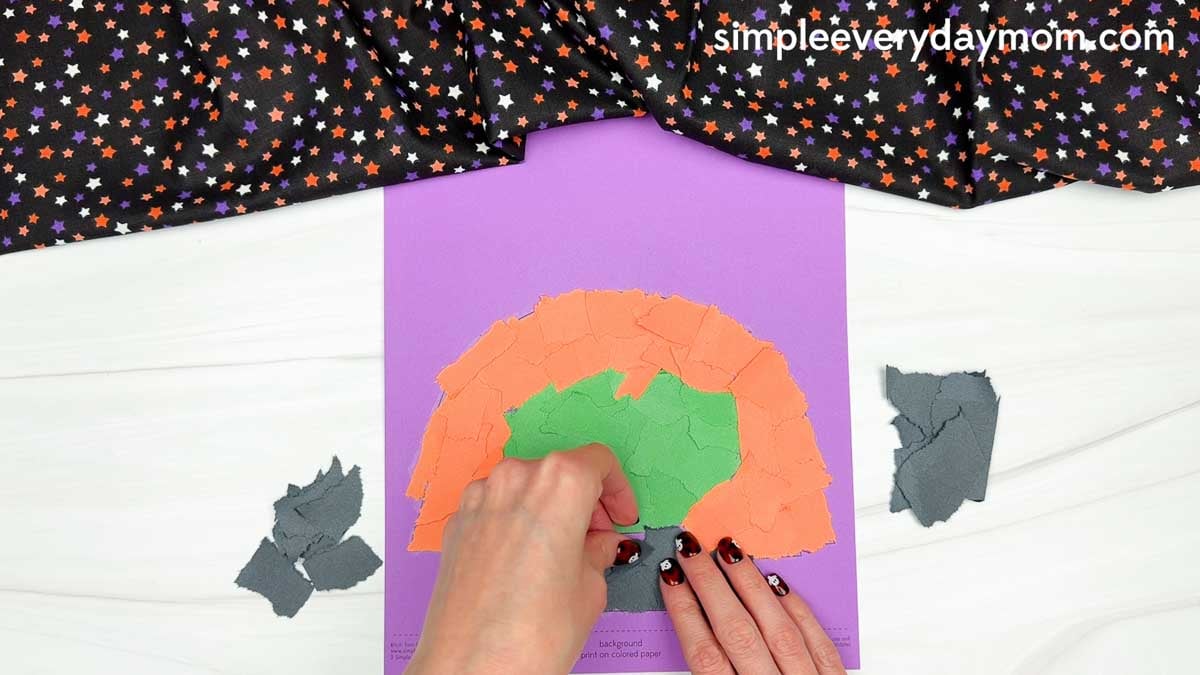 Hands gluing torn black paper pieces below the witch's green face to create the neck for the Witch Torn Paper Craft.
