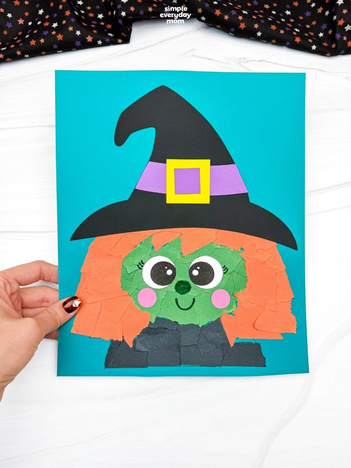 Hands holding a completed Witch Torn Paper Craft featuring a green face, orange torn paper hair, and a black witch hat on a blue background.