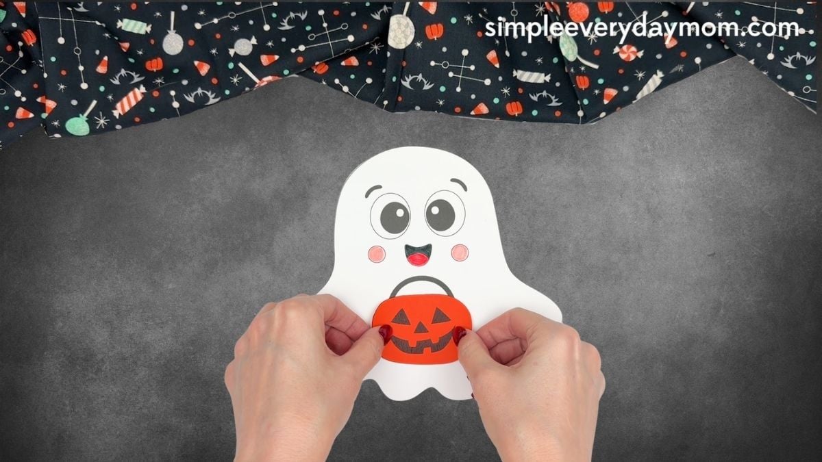 Hands placing an orange pumpkin bucket below the smiling face on a Halloween paper ghost craft.