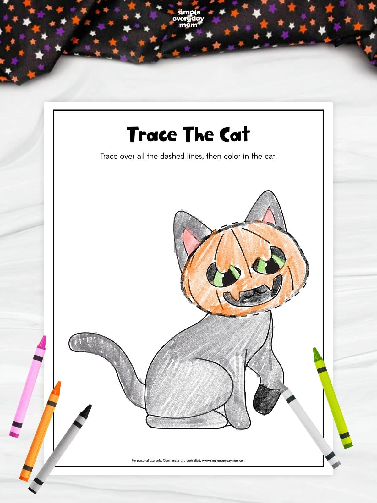 Cat tracing and coloring worksheet featuring a gray cat wearing an orange jack-o’-lantern mask with green eyes. The completed illustration is surrounded by pink, orange, gray, and green crayons.