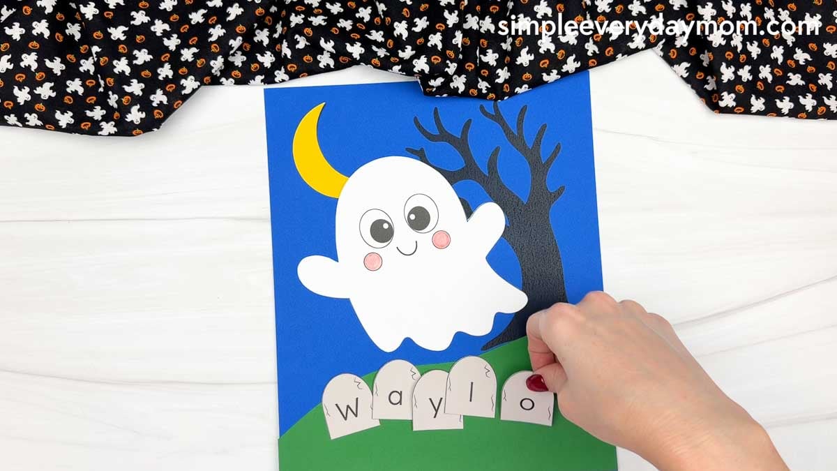 Hands arranging lettered tombstone pieces side by side on the green grass to spell a child's name for the Halloween Ghost Name Craft.