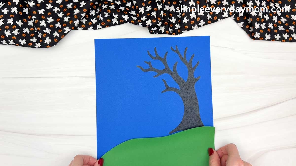 Hands positioning the green grass piece along the bottom of a blue background with a spooky black tree for the Ghost Name Craft.