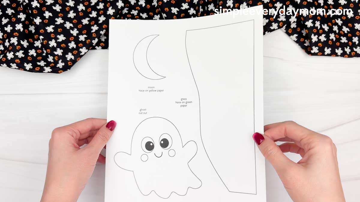 Hands holding a printable Ghost Name Craft template featuring a smiling ghost, crescent moon, and grass pieces for a Halloween paper activity.