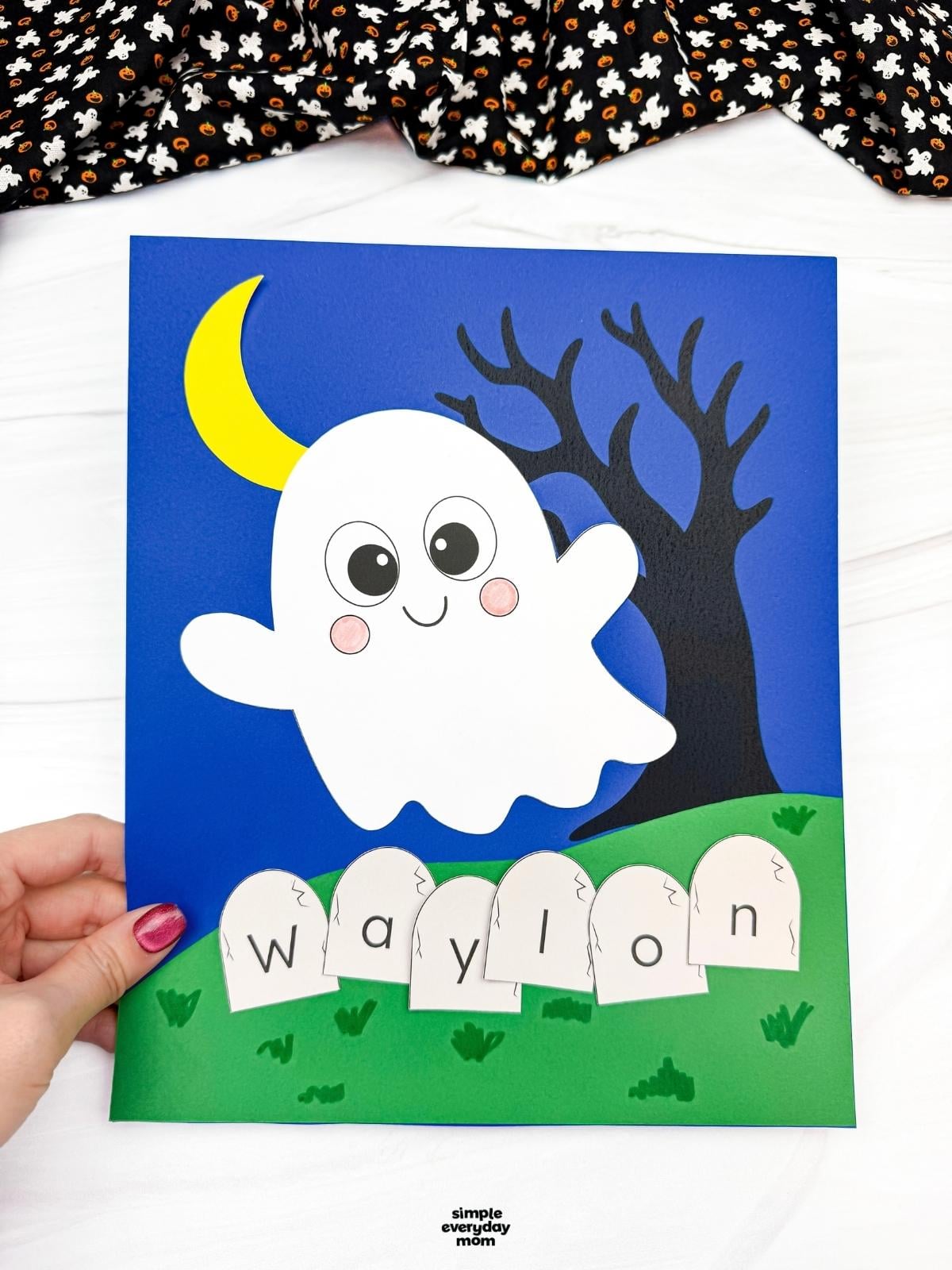 Hands holding a completed Ghost Name Craft featuring a smiling ghost, crescent moon, spooky tree, and lettered tombstones spelling a child's name.