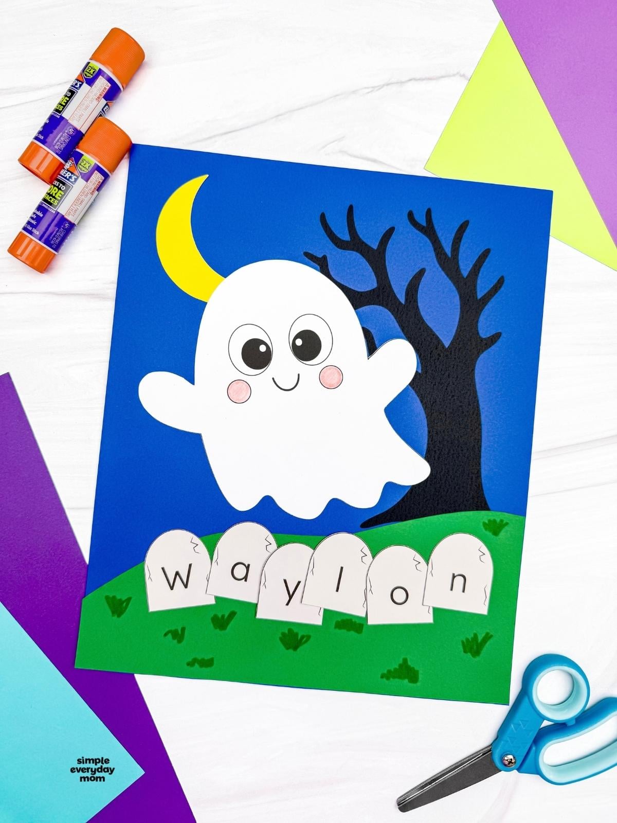 A completed Craft featuring a smiling ghost, crescent moon, spooky tree, and tombstones spelling a child's name, surrounded by colorful paper and craft supplies.