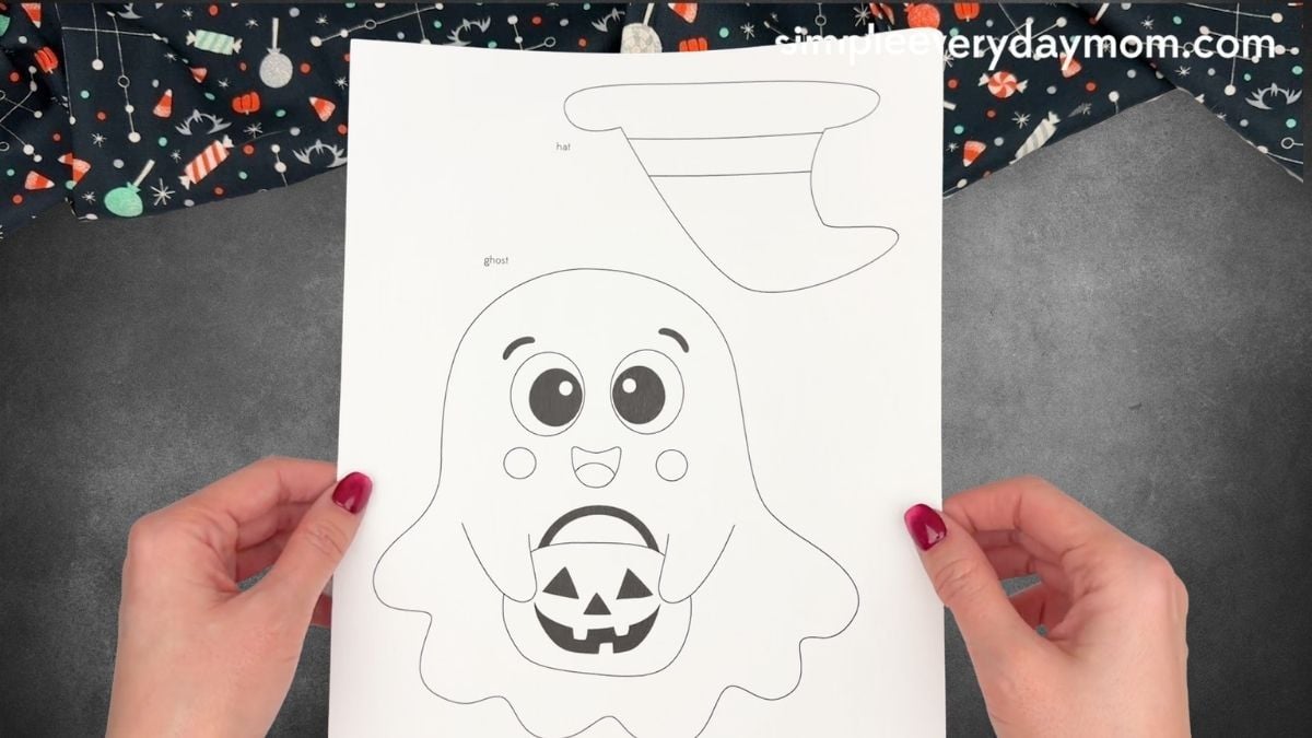 Hands holding a printable Halloween ghost craft template with a smiling ghost, pumpkin bucket, and witch hat.