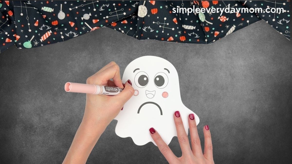 Hands coloring the cheeks pink on a Halloween paper ghost craft with a marker.
