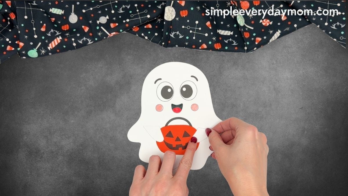 Hands attaching a white arm over the right edge of the pumpkin bucket on a Halloween paper ghost craft.
