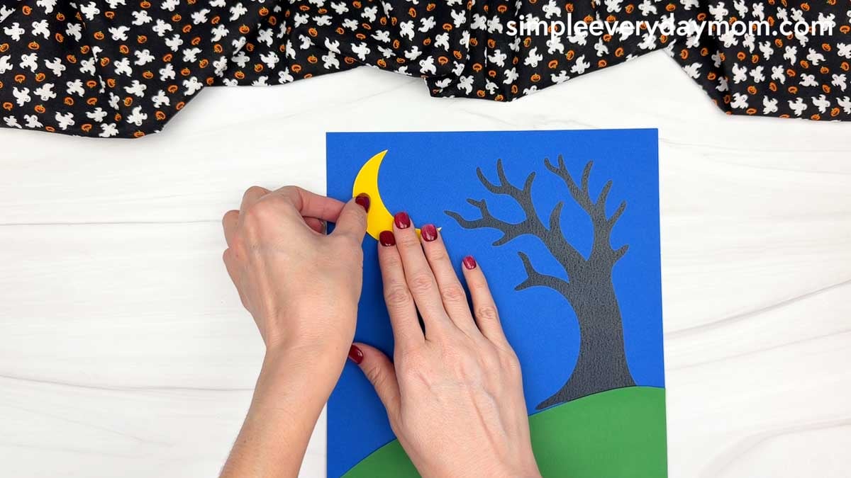Hands gluing the yellow crescent moon to the upper left of the blue background beside a spooky tree for the Ghost Name Craft.